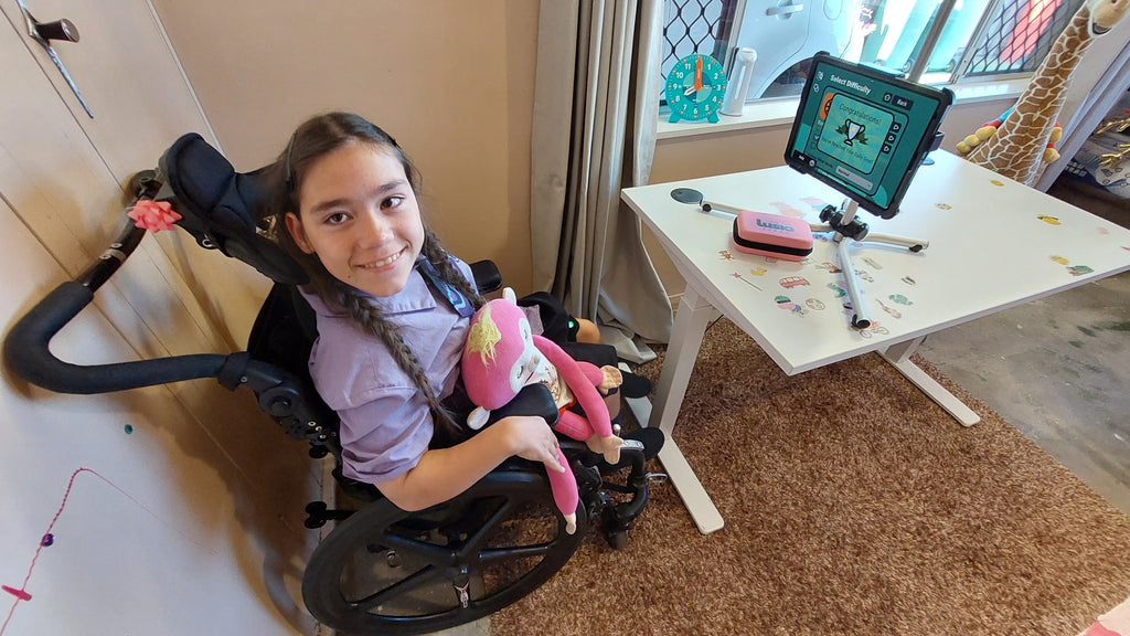 Emma Deede smiles brightly at the camera, radiating positivity in her wheelchair, with the engaging LusioMATE system visible in the background, showcasing the innovative physical therapy technology she's using.